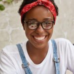 Headshot of pleasant looking young woman has shining smile, wears piercing in nose and lips, spectacles, red headband, being in high spirit after successfully completed task, content with result